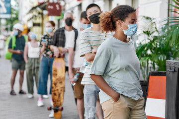 Young african american woman wearing mask waiting, standing in line with other people, respecting...