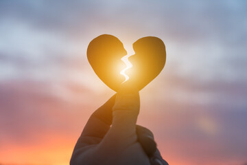 Silhouette of a hand holding a broken heart on the background of the sunset. Love concept, spat,...