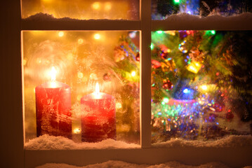 Night view from outside of a window decorated for Christmas. Lit candles, defocused Christmas tree and lights.