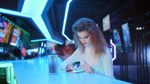 Trendy beautiful young woman in white costume use smartphone chatting in social media, typing and scrolling while sitting in night club bar backlit with neon light. Futuristic design, future world