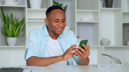Young mixed race man sitting at table, reading sms on smartphone. Focused millennial businessman checking email in mobile application, communicating with partners.