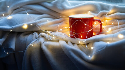 Christmas mood. Red mug of coffee or cocoa on white plaid with christmas lights. Homely Winter festive concept