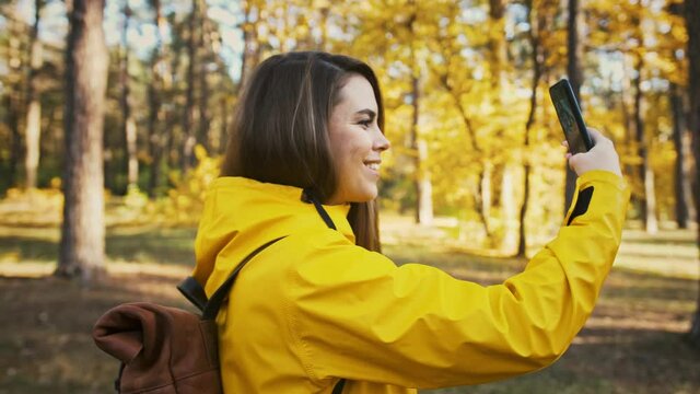 Young woman smiling, taking pictures of environment and selfies on her smartphone during walk in autumn wood