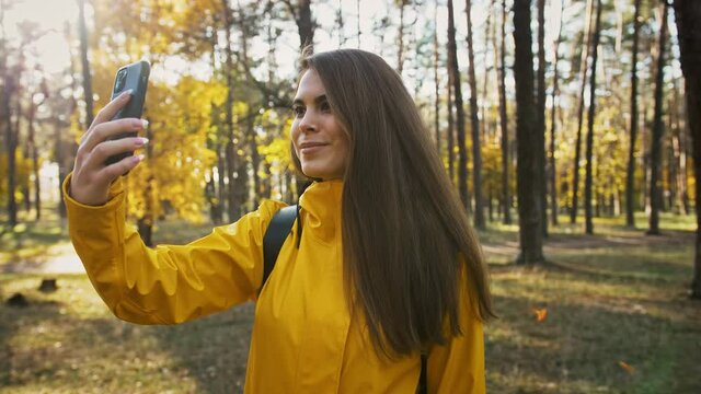 Young lady smiling, taking pictures of environment on her mobile phone during walk in autumn wood