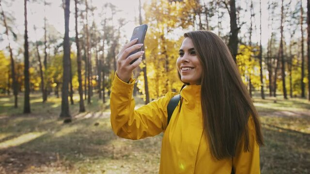 Young woman smiling, taking photos of environment and selfies on her smartphone during walk in autumn forest