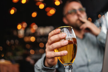Closeup of man holding a glass of fresh cold light beer, smoking cigarette and working on laptop.