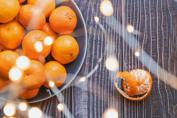 close up on a plate of ripe orange tangerines on a wooden table, one tangerine peeled, through Christmas lights bokeh - 395141195