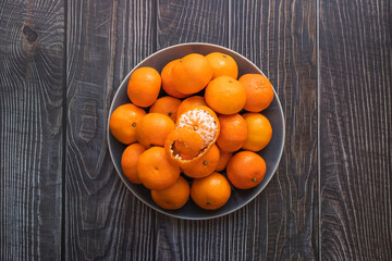 a plate of ripe orange tangerines on a wooden table, one peeled tangerine on top - 395141306