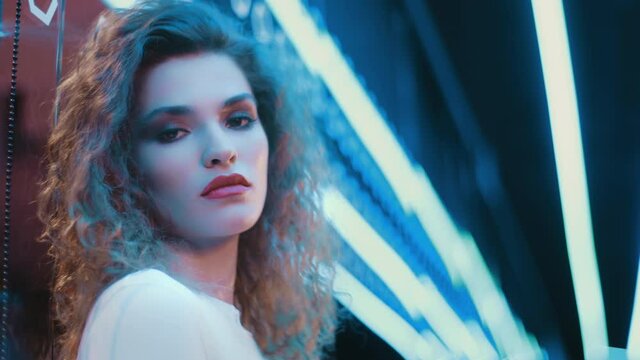 Portrait beautiful trendy woman wear white outfit posing looking at camera while standing by illuminated wall in night club. Cyberpunk futuristic style, sensual look clubber in blue violet neon lights