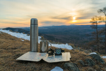 Terrain map compass thermos and a mug on a mountain top with sunset. Travel trip tourism concept. Navigation, route planning