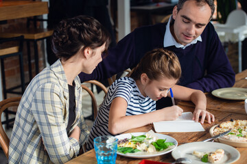 A girl doing her homework at the kitchen table with her parents being around.