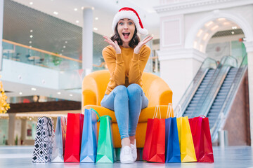 Excited young brunette woman in santa hat sitting in the mall with lots of colorful shopping bags