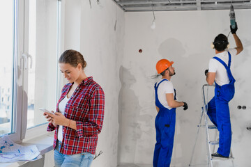A client in a plaid shirt and jeans uses the phone. A foreman in a construction helmet and his colleague are drilling a wall