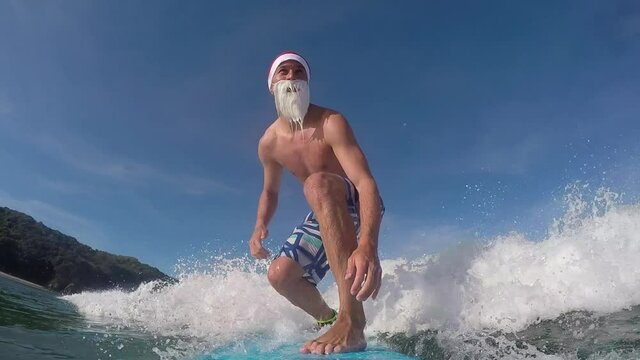 SLOW MOTION CLOSE UP: Cheerful Santa surfer riding fun waves in sunny ocean. Smiling surfer Santa Clause surfing waves towards the shore to wish a Merry Christmas. Funny santa Clause surfing