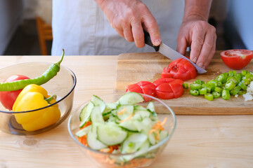 man in an apron prepares a salad of fresh vegetables at home, cuts paprika with a large knife, tomatoes, healthy food preparation concept, chopped ingredients, cucumbers, carrots