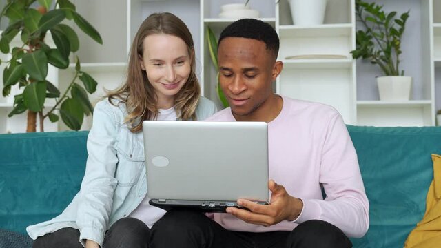 Cheerful interracial couple web surfing on laptop together, sitting on sofa at home