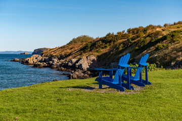 Two royal blue wooden Adirondack chairs on rich green grass overlooking the calm blue ocean under a...