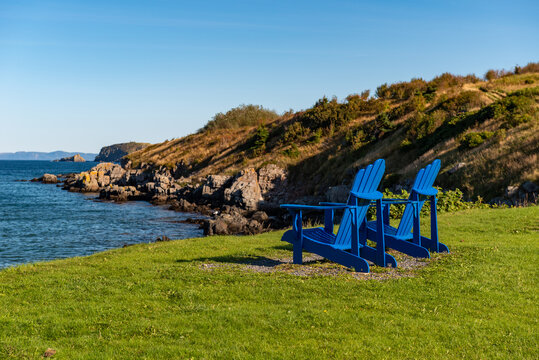 Two royal blue wooden Adirondack chairs on rich green grass overlooking the calm blue ocean under a bright blue sky.  There's a rocky shoreline in the distance with trees, shrubs and red foliage.