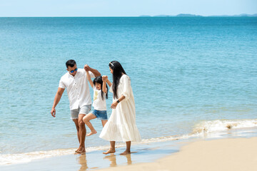 Happy Asian family parents with little daughter holding hand and walking together on the beach in summer day. Father and mother with cute child girl enjoy and having fun with outdoors holiday vacation