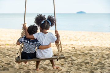 Happy adorable little mixed race girl with Asian girl friend in sitting on tree swing on island beach by the sea and hugging together. Cute child girl relax and having fun on summer holiday vacation.