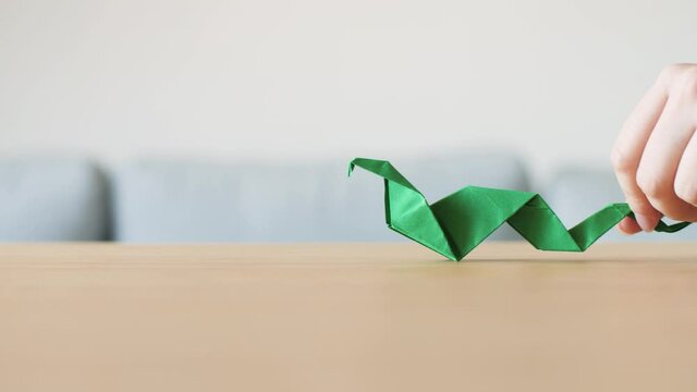 A hand moving a cute stylized green handmade Origami Snake on a wooden table. The animal was made with a traditional Japanese paper folding technique. Symbol for creativity, healing and wisdom. 4K.