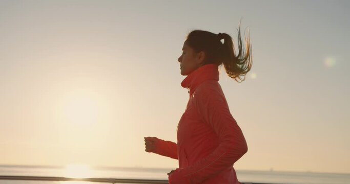 Young beautiful girl running in sports suit and with ponytail on background of sun rising on horizon over the sea.