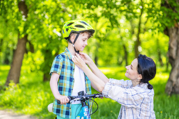 Naklejka na ściany i meble Mom hands out a bicycle helmet to a child standing with a bicycle in the summer in the park. Family Sports Vacation Concept