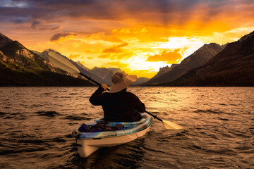 Naklejka na ściany i meble Adventurous Man Kayaking in Glacier Lake surrounded by the beautiful Canadian Rocky Mountains during a cloudy summer sunset. Taken in Upper Waterton Lake, Alberta, Canada.