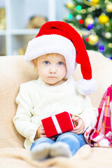 A little girl sits in a Santa hat at home on the sofa on a Christmas tree and holds a gift in her hands. Cozy holidays concept
