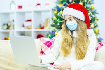 A girl in a santa hat and a medical mask waves a greeting to a laptop and congratulates online friends or relatives with merry christmas or new year during lockdown