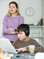 Son plaing video games at laptop, sad mom on background indoors