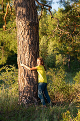 A girl with long hair and a yellow t-shirt hugs a large pine trunk in summer. The concept of unity with nature