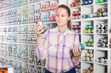 portrait of cheerful spanish woman choosing paint color in aerosol can in art shop