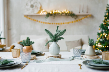 Stylish and trendy design of a festive table set for a family dinner. Vases with spruce branches, glasses and plates on the background of Christmas lights