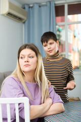 Offended mother sitting at table after quarrel with son in domestic interior