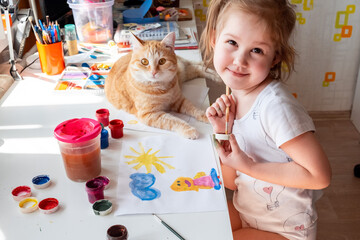 A little girl paints the sun and her mother with watercolors, a ginger cat lies next to the table. Children's creativity. Distance learning