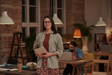Portrait of young female designer in eyeglasses holding tablet pc and smiling at camera standing at office