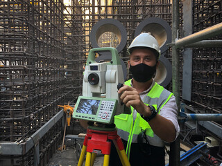 Land surveyor dressed in hardhat,mask and protective vest works on total station