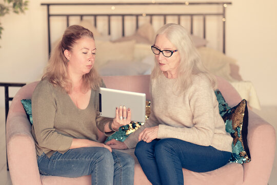 Woman teaching senior mother to use internet at home. Senior woman with her daughter looking at modern gadget indoors. relaxing together on couch indoors, different generations hobby pastime.