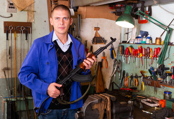 Portrait of confident master of weapons repair workshop standing near workplace with Kalashnikov...