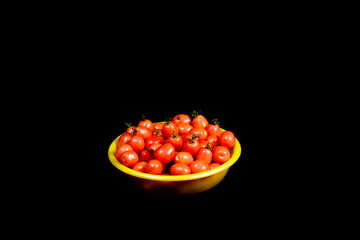 Harvest of fresh small tomatoes in a yellow plastic bowl on a black cloth background