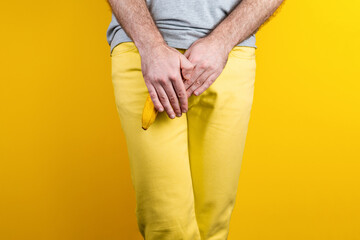 Impotence and men's health. A man in yellow jeans shyly covers a banana near the genitals with his hands. Yellow background. Close up of hands. Copy space