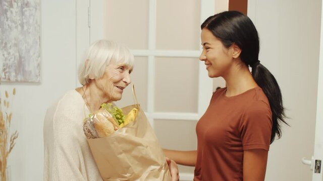 Young Asian female caregiver giving bag of groceries to cheerful senior woman and then posing for camera together with her at home