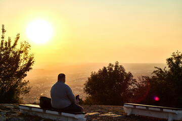 A big man sitting on a bench looking on a sunset