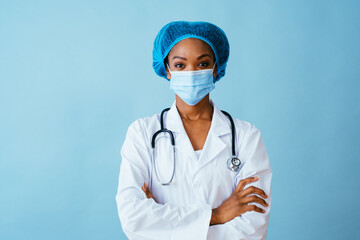 Portrait of a doctor in lab coat with face mask and hair protection with arms crossed, isolated on blue background