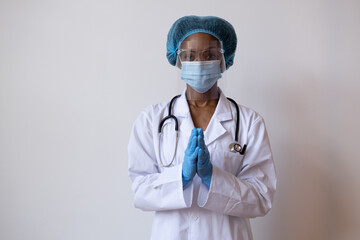 Portrait of a doctor in lab coat with face mask, face shield,  and hair ppe with gloved hands together as if saying please or praying