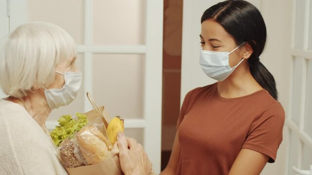 Young female volunteer in protective mask and gloves bringing bag of groceries to home of senior woman and speaking with her while helping pensioners during coronavirus quarantine