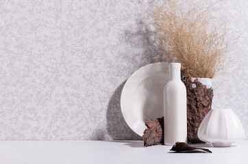 Scandinavian minimalist interior with ceramic vase, dish, dried grass bouquet, brown tree bark, moss in sun beam with shadow on white wood table.