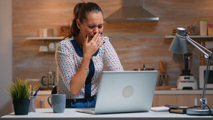 Woman yawning while working remotly from home late at night in the kitchen. Busy exhausted employee using modern technology network wireless doing overtime for job reading typing, searching