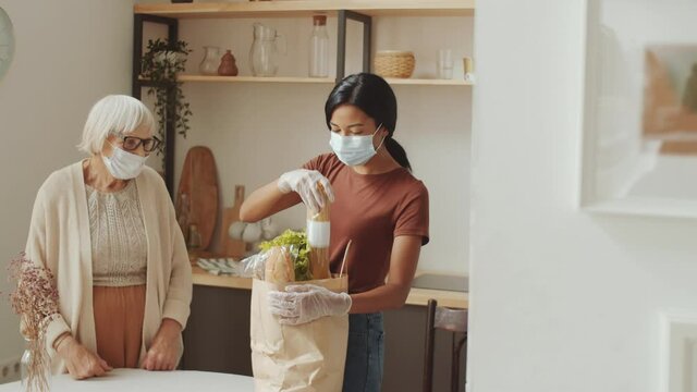 Young female caregiver in disposable mask and gloves unloading groceries from bag in the kitchen and speaking with senior woman while taking care of her during coronavirus quarantine